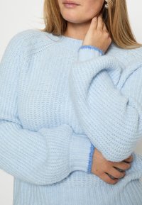 Gaiši zils adīts džemperis ar ribota raksta, ar garām piedurknēm un mīkstiem aprocēm, ko izceļ tumši zils akcenta tonis.