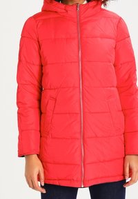 Červená zateplená bunda s kapucí, vybavená horizontálním prošíváním, předním zipem a bočními kapsami. Vyrobena ze syntetických materiálů, s hřejivou texturou.