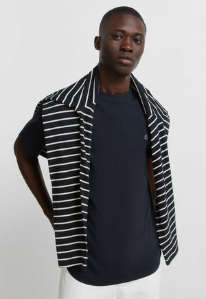 Jonge man die een marineblauw T-shirt draagt met een zwart-wit gestreepte trui over zijn schouders gedrapeerd, staand tegen een effen achtergrond.