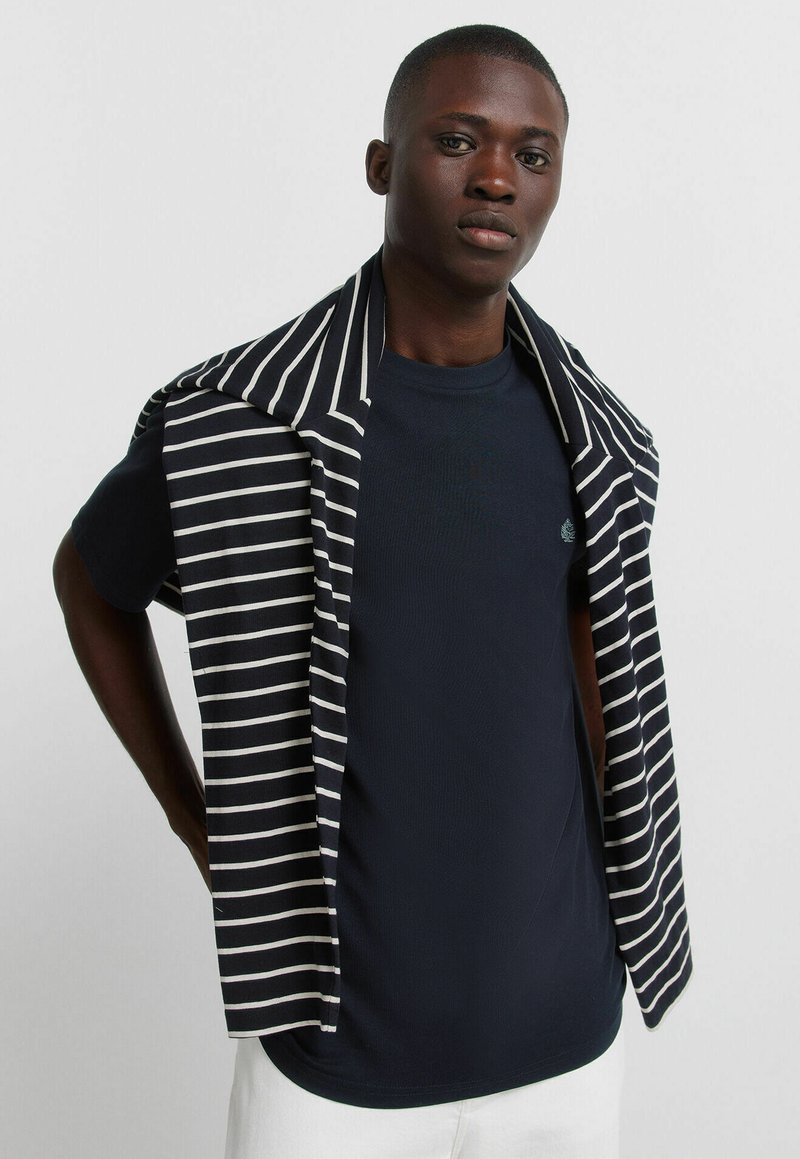 Młody mężczyzna w granatowej koszulce z czarno-białym swetrem w paski zarzuconym na ramiona, stojący na jednolitym tle.