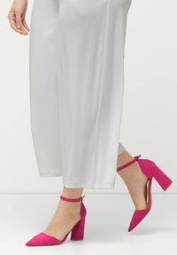 Fuchsia Wildleder-Pumps mit spitzem Absatz und Knöchelriemen, kombiniert mit fließenden weißen weiten Hosen.
