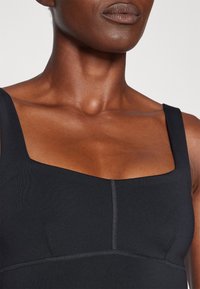 Μαύρη εφαρμοστή μπλούζα με τετράγωνη λαιμόκοψη, φαρδιές τιράντες και διακριτές ραφές. Η λεία υφή ενισχύει το μινιμαλιστικό σχέδιο.