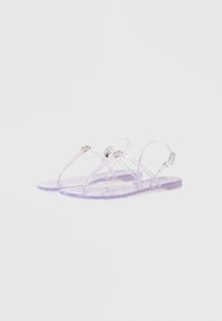 Gennemsigtige lilla sandaler med klare stropper og gulddetaljer, der har en flad sål og tekstureret fodseng. Justerbar ankestropp medfølger.