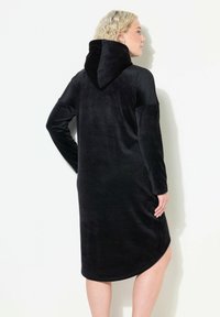 Schwarzes Langarmkleid aus weichem Stoff mit Kapuze, ausgestattet mit einem geschwungenen Saum und einem seitlichen Schlitz für mehr Bewegungsfreiheit.