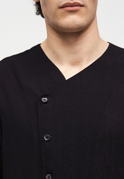 Άνδρας που φοράει μαύρο πουκάμισο με υφή και κουμπιά, με ασύμμετρο ντεκολτέ, ορατός από το πηγούνι έως το πάνω μέρος του στήθους σε λευκό φόντο.