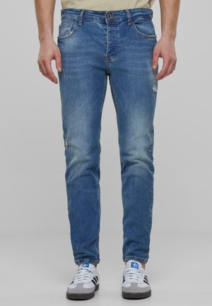 Jeans de denim azul con corte ajustado, zonas deslavadas y pequeño desgaste en los muslos. Combinados con zapatillas blancas y grises.