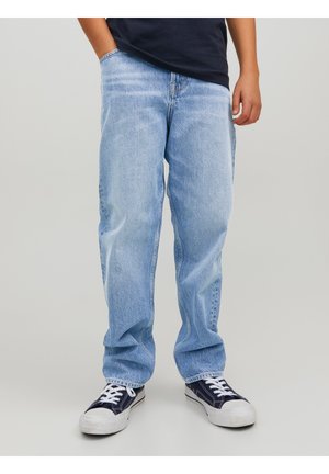 Barn iført lyseblå løse jeans og sorte lave sneakers med hvide snørebånd og såler, stående med den ene hånd i lommen.