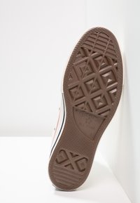 Semelle d'une basket blanche Converse All Star avec un motif de bande de roulement marron, appuyée contre un mur et un sol blancs.