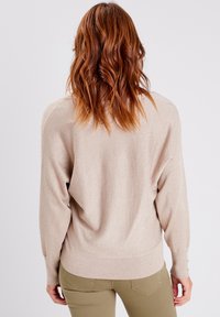 Pull oversized beige clair avec une texture scintillante, coupe lâche et ourlet côtelé ; dispose de manches longues et de détails en bouton aux poignets.
