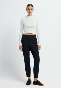 Witte geribbelde lange mouw crop top met een gedraaide voorkant, gedragen met marineblauwe tapered broeken met gouden knoopaccenten en bruine veterschoenen.