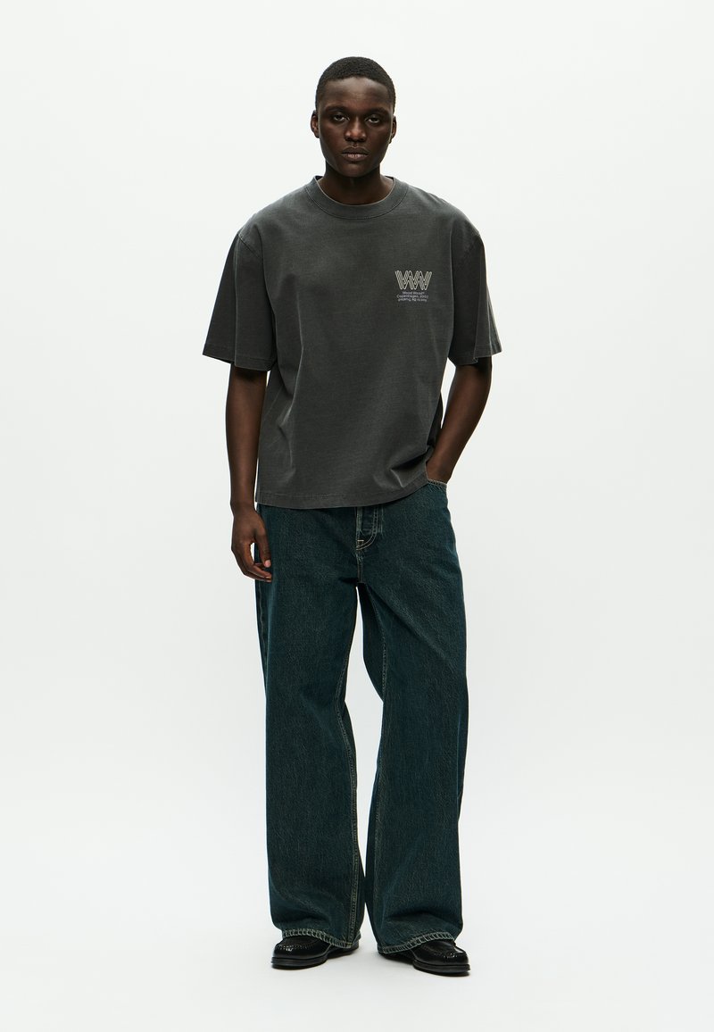 Young man wearing a loose dark gray logo T-shirt and wide-leg blue jeans, standing with one hand in pocket on a plain white background.