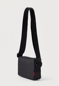 Μαύρη ορθογώνια τσάντα χιαστί με υφή, μαλακή κατασκευή και ρυθμιζόμενο λουράκι. Διαθέτει ένα μικρό κόκκινο λογότυπο ως διακόσμηση.