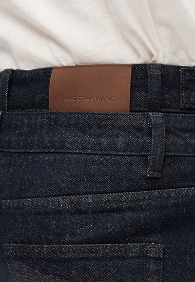 Σκούρο μπλε τζιν παντελόνι με περιοχή της μέσης στο πίσω μέρος, με καφέ δερμάτινη ετικέτα πάνω από την τσέπη και λευκό πουκάμισο που έχει μπει μέσα.