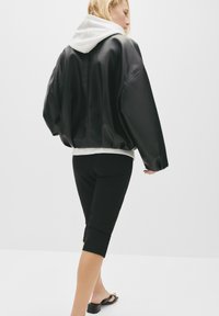Svart läder, oversized jacka med en grå huva, i kombination med svarta knälånga leggings och svarta sandaler. Slät textur, avslappnad silhuett.