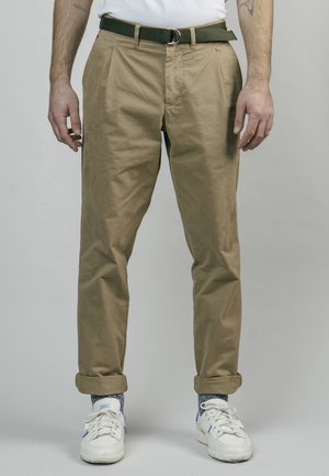 Homme portant un pantalon beige retroussé avec une ceinture noire, des baskets blanches et des chaussettes gris tachetées, debout devant un fond uni.