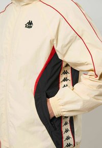 Lysebeige jakke med røde og sorte accenter, der har en broderet logo, elastiske manchetter og en sidelommer med lynlås. Stoffet virker glat.