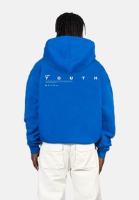 Blue hoodie made of thick fabric, featuring a large hood and white printed text on the back: "LOST YOUTH." White cargo pants are worn below.