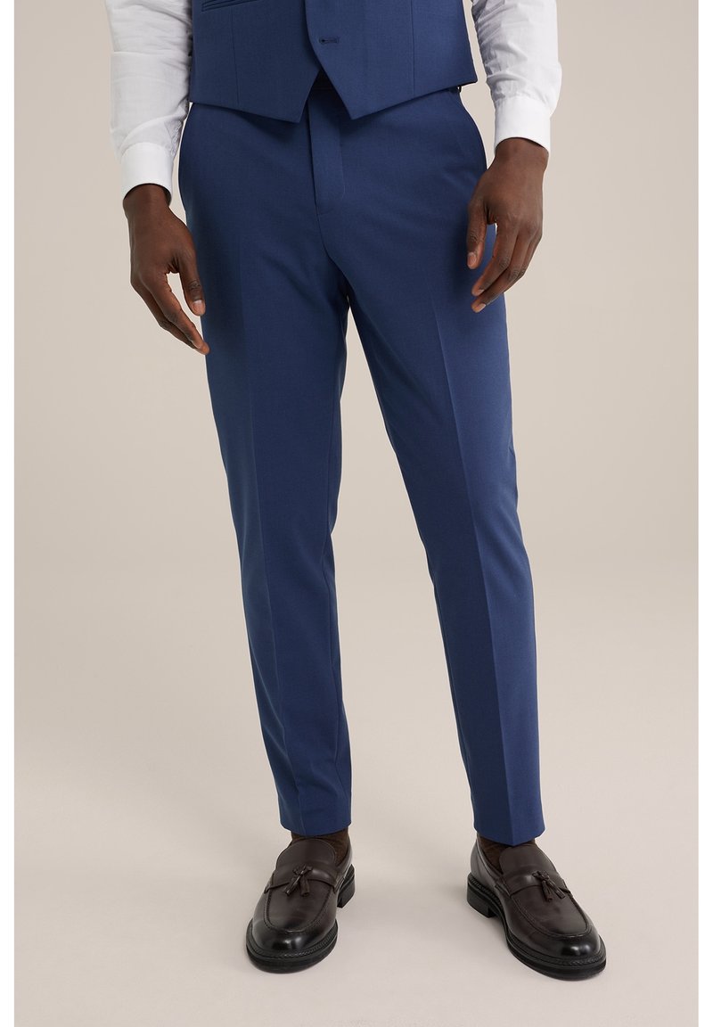 Homme portant un pantalon de costume bleu sur mesure, une chemise blanche, un gilet marine et des mocassins à pampilles marron foncé, debout devant un fond uni.