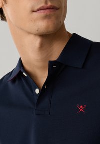 Mand iført marineblå polo med hvide knapper og lille rødt korslag af stok og hat-logo på brystet.