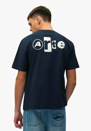 Giovane uomo di spalle che indossa una maglietta blu navy con la scritta "arte" e jeans blu con logo coordinato sulla tasca posteriore.