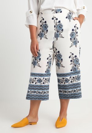 Mujer con pantalones blancos recortados con patrones florales azules y rayas, combinados con zapatos de punta amarillo mostaza sin cordones.