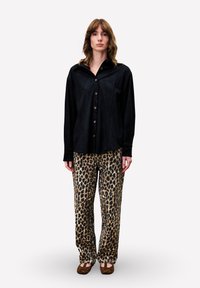 Eine Person trägt ein schwarzes Button-up-Shirt, weit geschnittene Hose mit Leopardenmuster und braune Ballerinas und steht vor einem schlichten weißen Hintergrund.