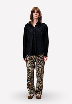 Persoon in een zwart overhemd met knopen, met een luipaardprint wijd uitlopende broek en bruine flats, staande tegen een eenvoudige witte achtergrond.