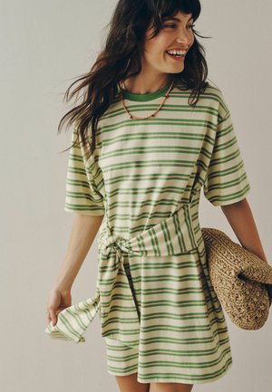 Femme souriante, portant une robe rayée vert et crème avec un nœud sur le côté, tenant une grande pochette tissée.