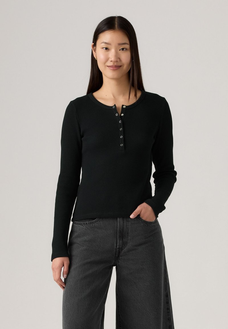 Sort langærmet henley-top lavet af ribbet stof, med en rund halsudskæring og fem sølvknapper. Bæres med mørkegrå bredbenede jeans.