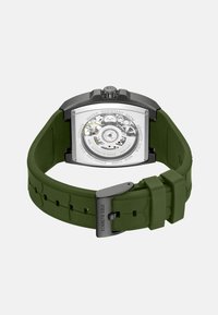 Quadratische Uhr mit transparentem Rücken, grünem Silikonarmband, Gehäuse aus Edelstahl, schwarzem Zubehör und sichtbaren mechanischen Komponenten.