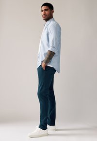 Lichtblauw gestreept overhemd met knopen, wit T-shirt eronder, donkergroene broek en witte sneakers. Model staat met hand in de zak.