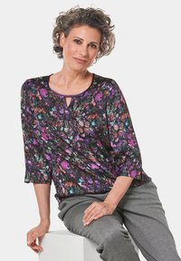 Mehrfarbige Blumenshirt mit Dreiviertelärmeln, Schlüssel-Ausschnitt und schwarzem Hintergrund. Getragen mit grauen gemusterten Hosen.