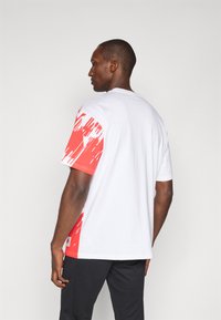 Hvid T-shirt med rød abstrakt print på venstre ærme og side. Klassisk crew neck-design i blødt stof, kortærmet.