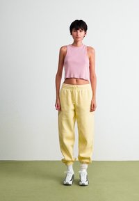 Kvinde står på grøn gulv iført en pink ærmeløs crop top, gule sweatpants, hvide sokker og sort-hvide sneakers.