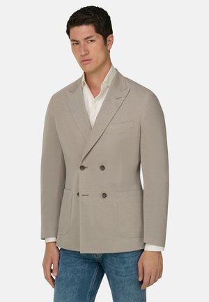 Uomo con blazer doppiopetto beige sopra camicia bianca e jeans blu, in piedi contro uno sfondo bianco semplice.