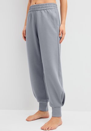 Persoon draagt losse, lichtgrijze joggingbroek met elastische tailleband en boorden, staat blootsvoets tegen een effen witte achtergrond.
