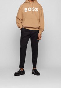 Persona vistiendo una sudadera color beige con el texto "BOSS", pantalones negros y mocasines negros, de pie contra un fondo gris claro liso.