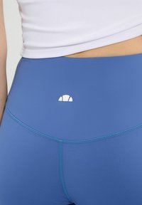 Niebieskie legginsy wykonane z elastycznego materiału, z białym logo z tyłu i kontrastowym przeszyciem wzdłuż szwów. Gładka tekstura.