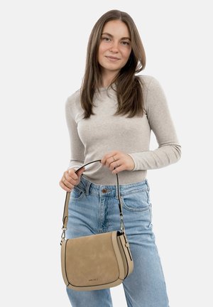 Sac à épaule en cuir beige avec une texture lisse, une forme courbée, une sangle réglable et des accents noirs. Le modèle tient le sac avec une tenue décontractée.