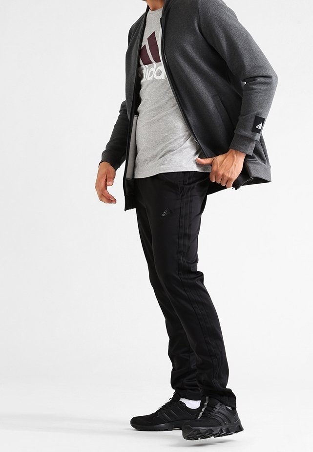 Chaqueta atlética gris con cremallera frontal, combinada con una camiseta gris que tiene un logo burdeos. Pantalones de chándal negros con rayas y zapatillas negras.