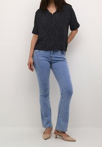 Blusa nera a maniche corte con pois bianchi e chiusura a bottoni, abbinata a jeans a zampa d'elefante blu chiaro e scarpe con tacco beige.