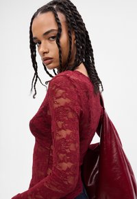 Top de renda bordeaux com mangas longas, apresentando padrões florais e um design ajustado, acompanhado de uma bolsa bordeaux brilhante. Textura suave e detalhes transparentes.