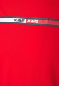 Punainen puuvillapaita, jossa on vaakasuuntaiset musta ja valkoinen raidat ja teksti "TOMMY JEANS" lihavoidulla mustalla ja punaisella fontilla. Sujuva pinta.