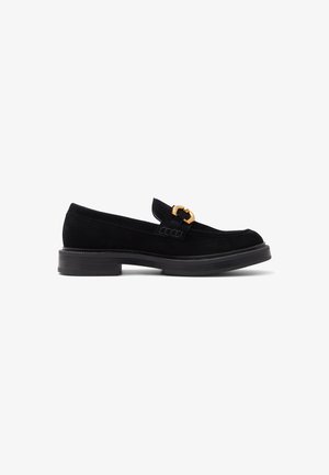 Mocassins en suède noir avec un accent de boucle en or, bout arrondi et une épaisse semelle en caoutchouc noir, présentant une texture lisse et un design raffiné.
