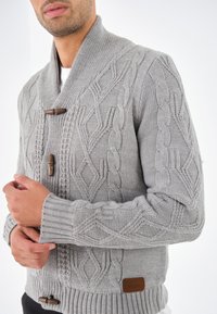 Grå stickad cardigan med hel krage, med flätstickat mönster, träspännen och ribbade manschetter och hem.