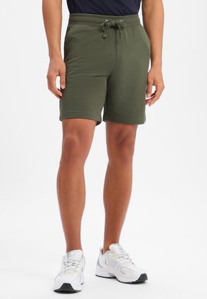 Mężczyzna w oliwkowo-zielonych szortach ze sznurkiem i białych sportowych trampkach, stojący na jednolitym białym tle.