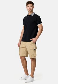 Crni polo majica s kontrastnim sivoj ovratnikom, uparena s bež cargo šortsevima koji imaju dva bočna džepa. Model nosi bijele tenisice.