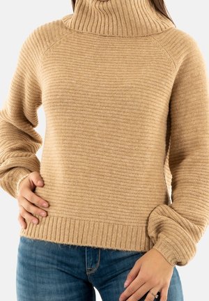 Femme portant un pull à col roulé côtelé beige à manches longues et un jean bleu, les mains posées près des hanches.