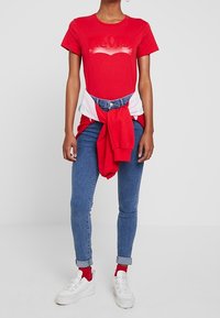 Rød bomulds T-shirt med et falmet Levi's logo, parret med blå skinny jeans, hvide sneakers og en rød sweater bundet om taljen.