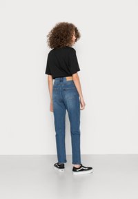 Jeans azuis de cintura alta com corte reto, bolsos traseiros, e um patch de pele castanha na cinta, combinados com uma T-shirt preta e ténis pretos.